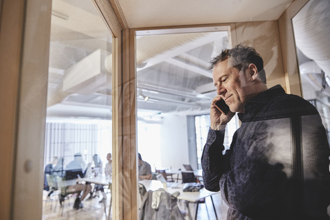 Älterer Geschäftsmann, der über sein Smartphone spricht, gesehen von einem Glas im Büro, lizenzfreies Stockfoto