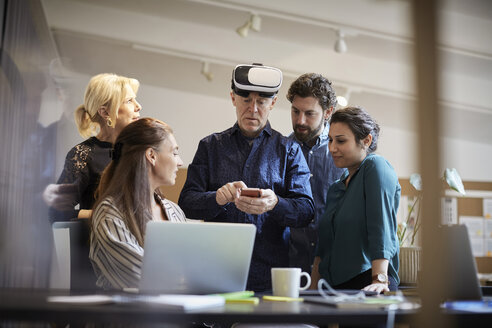 Mature businessman with virtual reality simulator using smart phone while standing amidst colleagues - MASF01653