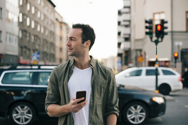 Man looking away while holding smart phone in city - MASF01657