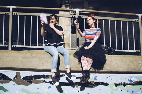 Hipster Freundinnen nehmen Selfie mit Handys, während sie an der Wand gegen Geländer sitzen, lizenzfreies Stockfoto