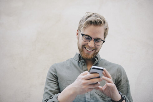 Glücklicher männlicher Computerprogrammierer, der ein Smartphone an einer beigen Wand im Büro benutzt - MASF01780