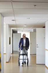 Älterer Mann in voller Länge, der wegschaut, während er mit einem Rollator im Krankenhausflur geht - MASF01782