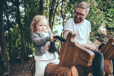 Glücklicher Vater sieht seine Tochter auf einem Holzpferd im Park reiten - MASF01789