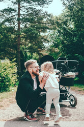 Glücklicher Mann hockt auf dem Gehweg im Park und hält seine Tochter am Kinderwagen - MASF01790