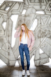 Full length portrait of smiling blond girl showing peace sign while standing against concrete wall at parking garage - MASF01831