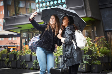 Glückliche Freunde mit Regenschirm, die ein Selfie mit ihrem Mobiltelefon machen, während sie in der Stadt stehen - MASF01842