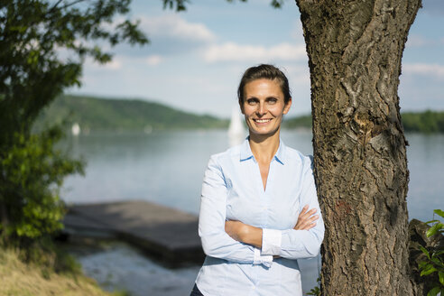 Porträt einer lächelnden Frau, die sich an einen Baum an einem See lehnt - JOSF02169