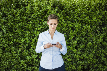 Woman standing at a hedge using cell phone - JOSF02170