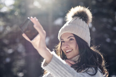 Porträt einer lächelnden jungen Frau, die ein Selfie mit ihrem Smartphone im Winterwald macht - ABIF00300