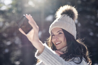 Portrait of smiling young woman taking selfie with smartphone in winter forest - ABIF00300