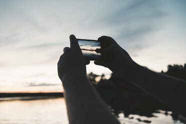 Cropped image of man photographing lake through smart phone during sunset - MASF01867