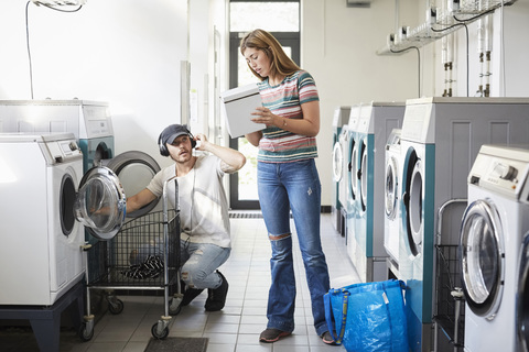 Verwirrte Studentin zeigt Waschmittelpackung an männlichen Freund im Waschsalon, lizenzfreies Stockfoto