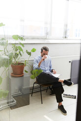 Geschäftsmann in voller Länge, der Kaffee trinkt und neben einer Topfpflanze im Büro ein Dokument liest - MASF01897