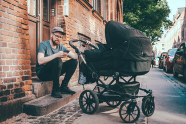 Vater, der sein Smartphone benutzt, während er den Kinderwagen auf den Stufen am Gebäudeeingang hält - MASF01898