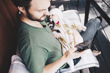 Father with toddler using mobile phone while sitting on chair at holiday villa - MASF01911