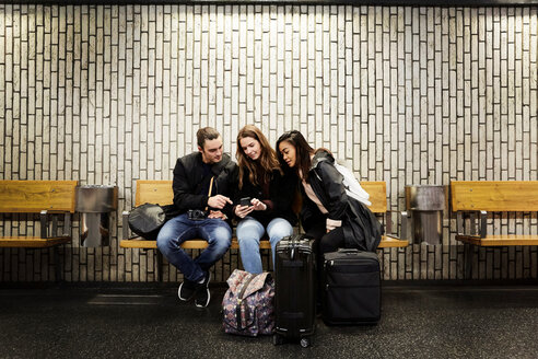 Freunde schauen in voller Länge auf ihr Smartphone, während sie auf dem Bahnsteig einer U-Bahn-Station an der Wand sitzen - MASF01925