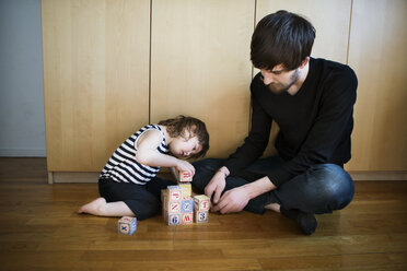 Father looking at daughter playing with toys blocks while sitting on floor - CAVF35398