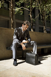 Man with briefcase looking at wristwatch while sitting on bench against building - CAVF35400