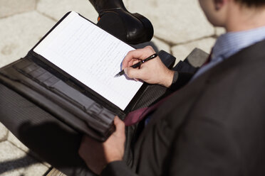 Hoher Blickwinkel eines Geschäftsmannes, der auf einer Bank sitzend an einem Dokument schreibt - CAVF35402