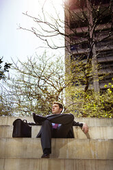 Geschäftsmann schaut weg, während er auf einer Stützmauer vor einem Gebäude sitzt - CAVF35404