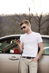 Man with hand in pocket using smart phone while leaning on car - CAVF35414