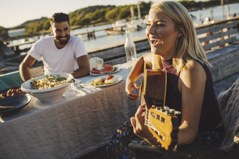 Fröhliche Frau spielt Gitarre, während ihr Freund an einem sonnigen Tag am Picknicktisch sitzt, lizenzfreies Stockfoto