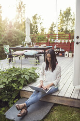 Frau, die einen Laptop benutzt, während sie auf einer Veranda im Hinterhof sitzt - MASF01950