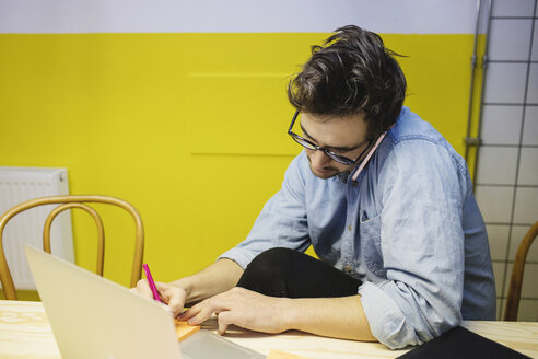 Junger Mann benutzt sein Smartphone und schreibt auf einem Klebezettel am Schreibtisch im Sitzungssaal - MASF01963