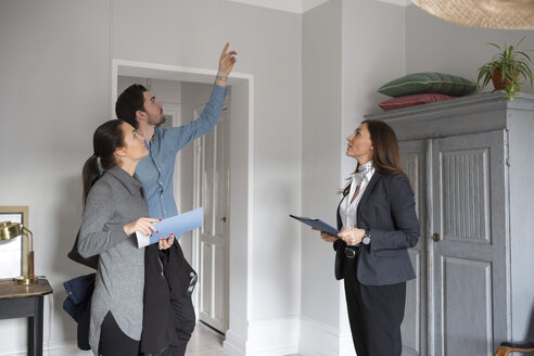 Frau und weiblicher Immobilienmakler schauen auf einen Mann, der nach oben zeigt, während er im Raum steht - MASF01986
