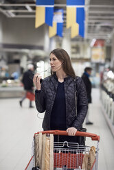 Frau spricht über Kopfhörer mit einem Mobiltelefon, während sie mit einem Einkaufswagen im Supermarkt steht - MASF01998