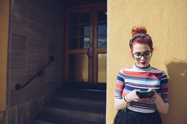 Junger weiblicher Hipster, der ein Mobiltelefon benutzt, während er an der Wand vor dem Eingang eines Gebäudes steht - MASF02002
