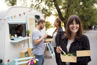 Smiling customer holding disposable salad box on street with friends and owner outside food truck in background - MASF02018