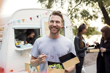 Smiling male customer holding disposable salad box against food truck with friends and owner in background - MASF02019