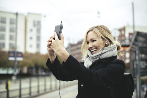 Lächelnde Geschäftsfrau im mittleren Erwachsenenalter, die an einer Straßenbahnhaltestelle ein Selfie mit ihrem Mobiltelefon macht - MASF02032
