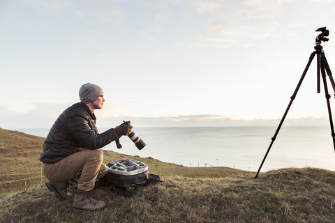 Seitenansicht eines Wanderers, der wegschaut, während er eine Spiegelreflexkamera auf einem Hügel am Meer gegen den Himmel hält, lizenzfreies Stockfoto