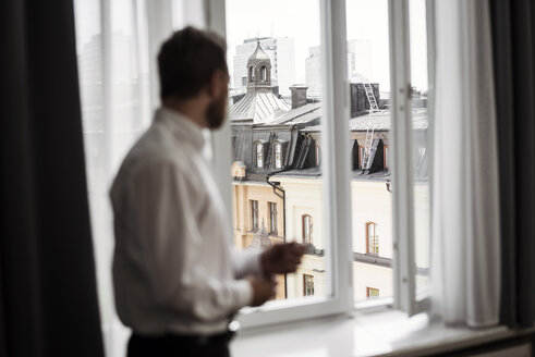 Seitenansicht eines Geschäftsmannes, der vom Hotelzimmer aus durch ein Fenster auf Gebäude blickt - MASF02084