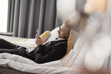 Seitenansicht eines Geschäftsmannes, der Musik hört, während er auf dem Bett eines Hotelzimmers liegt - MASF02086