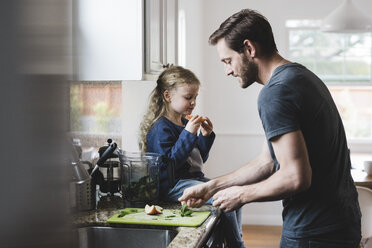 Seitenansicht eines Vaters, der Essen kocht, während seine Tochter in der Küche einen Apfel isst - MASF02104