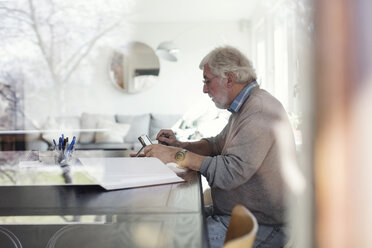 Seitenansicht eines älteren Mannes, der ein digitales Tablet benutzt, während er Papierkram erledigt, gesehen durch ein Glas - MASF02107