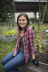 Porträt einer lächelnden erwachsenen Frau, die auf dem Rand einer Holzkiste im städtischen Garten sitzt - MASF02130