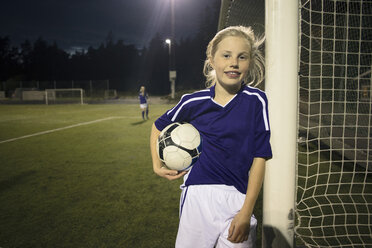 Porträt eines glücklichen Mädchens, das einen Fußball hält und am Torpfosten auf einem Feld steht - MASF02149