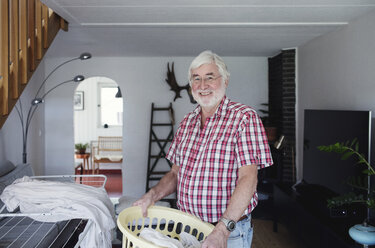Portrait of smiling senior man holding laundry basket while standing in living room - MASF02155