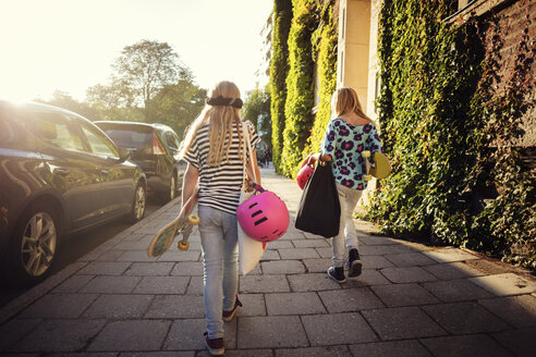 Rückansicht von Mädchen mit Skateboards, die an einem sonnigen Tag auf dem Gehweg neben geparkten Autos laufen - MASF02162