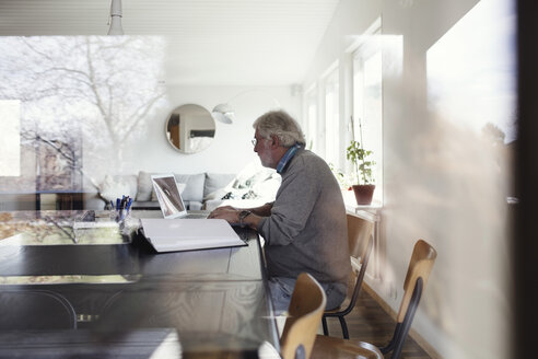 Älterer Mann, der einen Laptop benutzt, während er am Tisch sitzt, gesehen durch ein Fenster - MASF02190