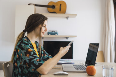 Young woman sitting at table at home using cell phone and laptop - KKAF00912