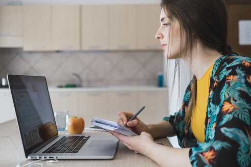 Young woman sitting at table at home using laptop and taking notes - KKAF00917