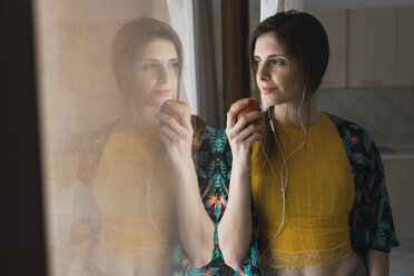 Junge Frau mit Kopfhörern und Apfel schaut aus dem Fenster - KKAF00922
