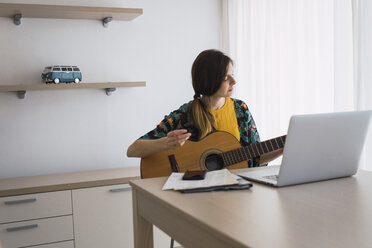 Young woman sitting at table at home with laptop playing guitar - KKAF00926