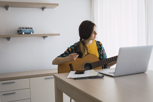 Junge Frau sitzt am Tisch zu Hause mit Laptop und spielt Gitarre - KKAF00926