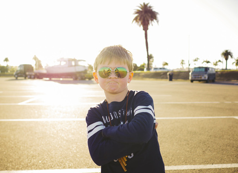 Porträt eines selbstbewussten Jungen mit Sonnenbrille und verschränkten Armen auf einem Parkplatz an einem sonnigen Tag, lizenzfreies Stockfoto